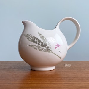 May include: A white ceramic pitcher with a rounded body and a curved handle. The pitcher is decorated with a gray and white leaf design and a small pink flower. The pitcher sits on a wooden surface.