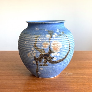 May include: A round ceramic vase with a textured, ribbed surface in shades of blue. The exterior features a mottled glaze with white and brown crystalline patterns and dark, organic-looking markings that wrap around the center of the vessel.