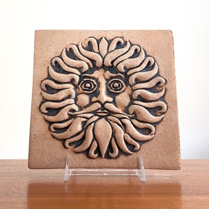 May include: A square, tan ceramic tile featuring a detailed, relief-style face with a long beard and flowing hair. The face has large, circular eyes and a prominent nose. The tile is displayed on a clear acrylic stand.