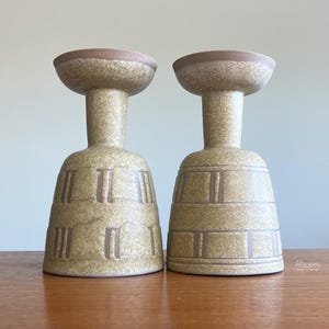 May include: Two matching ceramic candle holders. The candle holders are beige with brown accents and a textured surface. The base is a bell shape with vertical and horizontal brown lines. The top is a shallow bowl shape.