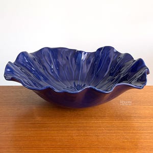 May include: A large, deep blue ceramic bowl with a ruffled, petal-like edge. The bowl has a glossy finish and sits on a wooden surface. The design is reminiscent of a flower or a sea creature.