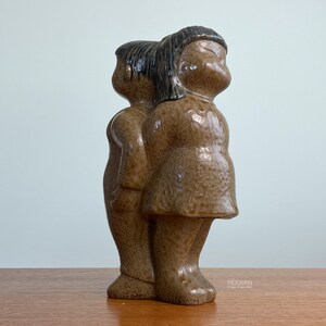 May include: A ceramic figurine featuring two figures standing back-to-back, finished in a glossy, speckled brown glaze. The figures have dark brown hair and are sculpted with smooth, rounded features, capturing a unified, joined composition.