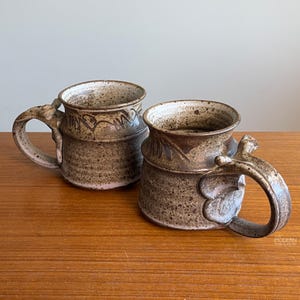 May include: Two handcrafted ceramic mugs with a speckled brown glaze. Each mug features a unique handle design and decorative detailing around the rim. The mugs are displayed on a wooden surface.