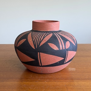 May include: A terracotta-colored ceramic vase with a rounded body and a short neck. The vase is adorned with a black geometric pattern featuring stylized leaf and triangle shapes. The vase sits on a wooden surface.
