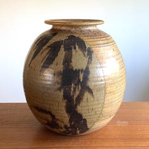 May include: A round ceramic vase with a wide body and a narrow neck. The surface features a textured, tan and beige base with horizontal ridges and prominent, dark brown, abstract brushstroke patterns. The rim is flat and slightly flared.