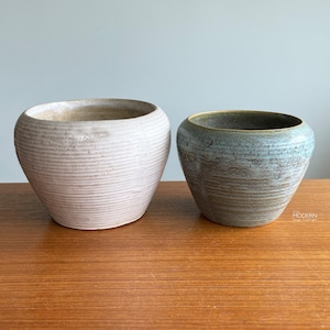 May include: Two ceramic planters with a textured, ribbed design. One is light pink, the other is blue-green with a darker rim. Both have a rounded, bowl-like shape and sit on a wooden surface. The planters are suitable for indoor or outdoor use.