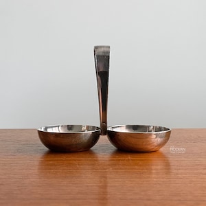 May include: A silver-toned condiment server with two small, round bowls connected by a handle. The bowls are side-by-side, and the handle extends upwards. The server is on a wooden surface.