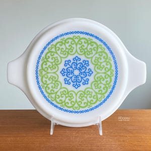 May include: White Pyrex lid with a decorative pattern in blue and green. The design features a central blue snowflake-like shape surrounded by green and blue swirls. The lid has two handles and is displayed on a clear stand.