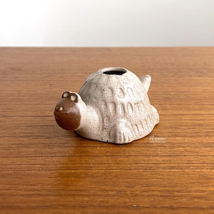 May include: A small ceramic turtle figurine with a speckled off-white shell and a smooth brown head. The shell features a textured pattern and a central circular opening at the top. The text "AJA MODERN" is printed in small letters on the side.