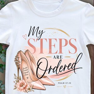 May include: A white short-sleeve t-shirt featuring a graphic of two light pink high-heeled shoes with gold accents. The text reads My STEPS ARE Ordered and Psalm 37:23, with additional small text inside the shoe and floral embellishments.