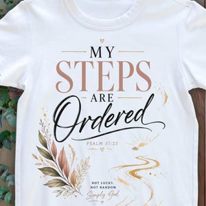 May include: A white short-sleeved t-shirt featuring the text MY STEPS ARE Ordered and PSALM 37:23. Below this, the text reads NOT LUCKY. NOT RANDOM. Simply God. The design includes a botanical illustration of leaves and a winding path graphic.