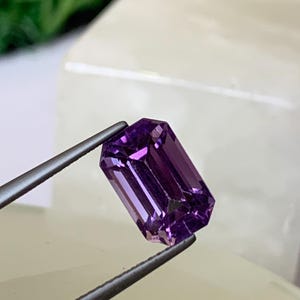 May include: A rectangular-cut amethyst gemstone, held by tweezers. The gem is a deep purple colour with internal facets that reflect light. The background is a soft, neutral tone, highlighting the gem's brilliance.