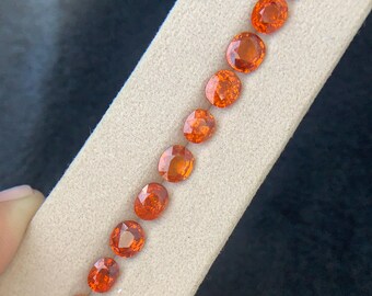 May include: A collection of vibrant orange gemstones, varying in shape from oval to round, are arranged on a light beige surface. The gemstones exhibit a brilliant, faceted cut, reflecting light. The background is a dark, neutral tone, highlighting the gems.