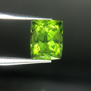 Könnte beinhalten: Ein leuchtend grüner, quadratisch geschliffener Peridot-Edelstein, der von einer Pinzette vor dunklem Hintergrund gehalten wird. Die Facetten des Edelsteins reflektieren das Licht und zeigen seine leuchtend limettengrüne Farbe. Dieser Edelstein ist ein schönes Beispiel für Peridot, eine beliebte Wahl für Schmuck.