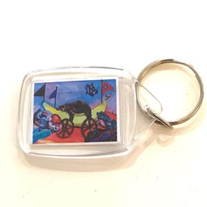 Puede incluir: Un llavero de plástico transparente con una imagen colorida de un oso negro tirando de un carro con ruedas. El carro está lleno de plátanos y el oso lleva un sombrero rojo. Hay mariposas azules y banderas en el fondo.