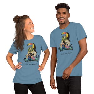 May include: Two people are wearing light blue t-shirts with a colorful illustration of a toucan, a frog, and two people in a bed. The text "Artist: Adam Mayfield" is printed on the shirts.