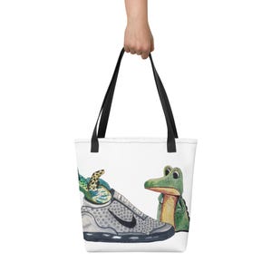 May include: A white tote bag with a black handle features a watercolor illustration of a grey sneaker with a green and yellow lizard inside and a green alligator looking at the sneaker.