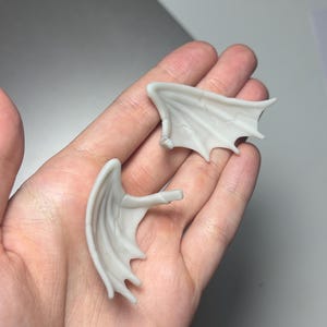 May include: Two small, light gray dragon wings with detailed vein patterns. Each wing has a curved shape with pointed tips and a small peg extending from the base. The wings are designed for crafting or miniature projects.
