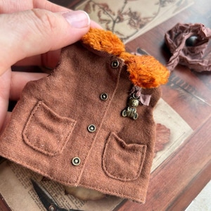 Op de afbeelding: Een miniatuur bruin vest met een oranje bontkraag, twee voorzakken en drie messing knopen. Een kleine teddybeer hanger hangt aan het vest. Het vest wordt door een hand vastgehouden en rust op een vintage boek met een vogelillustratie.