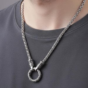Può includere: Collana a catena color argento con un ciondolo circolare. La collana presenta un design a catena intrecciata e un ciondolo ad anello decorativo. Il gioiello è indossato da una persona che indossa una maglietta grigia.