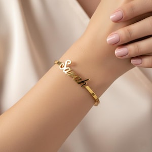May include: A gold-coloured bangle bracelet inscribed with the name "Sarah" in a cursive font. The bracelet has a simple, elegant design, with the name as the main feature. The bracelet is worn on a wrist.