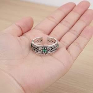 Könnte beinhalten: Verstellbarer Ring in Silberoptik mit keltischem Knotenmuster und grünen Edelsteinakzenten. Der Ring hat ein offenes Design und wird auf einer Hand gezeigt, um seine komplizierten Details und seine Handwerkskunst hervorzuheben. Der Ring ist ein Schmuckstück.