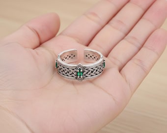 May include: Silver-toned adjustable ring with a Celtic knot design and green gemstone accents. The ring features an open design and is displayed on a hand, showcasing its intricate details and craftsmanship. The ring is a piece of jewelry.