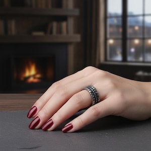Può includere: Un anello d'argento con un design geometrico è indossato su una mano con smalto bordeaux. L'anello ha una superficie strutturata e un aspetto impilato. Lo sfondo include un camino e una finestra.