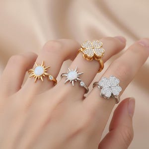Könnte beinhalten: Vier Ringe werden gezeigt, jeder mit einem einzigartigen Design. Zwei Ringe haben ein Sonnenmuster mit einem zentralen opalartigen Stein, einer goldfarben und einer silberfarben. Die anderen beiden Ringe haben ein vierblättriges Kleeblatt-Design, eines goldfarben und eines silberfarben, beide mit kleinen, funkelnden Steinen besetzt.
