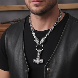 May include: A silver-colored necklace featuring a Thor's hammer pendant. The necklace has a chain design with wolf head accents. The pendant is detailed with engraved patterns. The necklace is worn over a black shirt and a black leather vest.