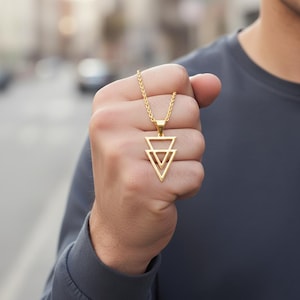 Op de afbeelding: Goudkleurige ketting met een hanger met twee overlappende driehoeken. De ketting en hanger hebben een glanzende afwerking. De ketting wordt tegen een vage achtergrond gehouden, wat wijst op een buitenomgeving. De sieraden zijn eenvoudig en geometrisch.