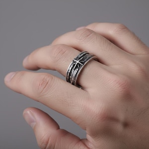 May include: A silver-toned ring with a star design. The ring features a textured band with a central star motif. The ring is worn on a finger, showcasing its intricate details and metallic sheen.