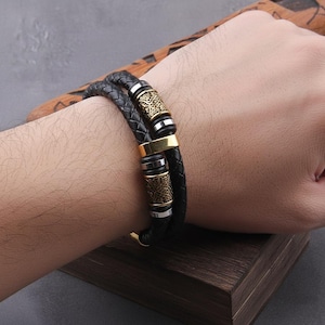 Puede incluir: Pulsera de cuero trenzado negro con detalles dorados y plateados. La pulsera presenta cilindros metálicos decorativos con intrincados patrones. La pulsera se lleva en la muñeca.