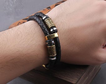 May include: Black braided leather bracelet with gold and silver accents. The bracelet features decorative metal cylinders with intricate patterns. The bracelet is worn on a wrist.
