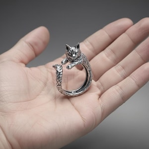 May include: A silver-colored cat ring. The ring features a detailed cat design with the cat's head and paws forming the top of the ring, and the tail curving around the finger. The ring is made of a shiny metal and is set against a neutral background.