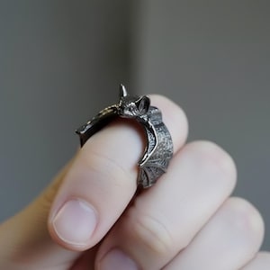 Puede incluir: Un anillo de murciélago gris oscuro. El anillo presenta un diseño detallado de murciélago con alas extendidas y una superficie texturizada. La cabeza del murciélago es visible en la parte superior del anillo, con sus orejas y cara claramente definidas. El anillo se lleva en un dedo.