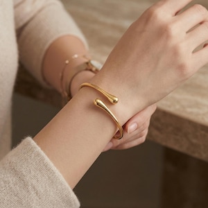 Könnte beinhalten: Ein goldfarbenes Armreif-Armband mit tropfenförmigen Enden. Das offene Design ermöglicht ein einfaches Tragen. Das Armband wird an einem Handgelenk gezeigt, was auf ein Schmuckthema hindeutet.