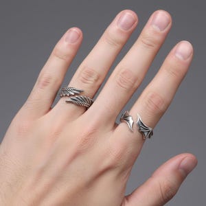 Puede incluir: Dos anillos plateados con diseños de alas. Un anillo presenta un diseño de ala detallado, mientras que el otro tiene un diseño de ala de murciélago. Los anillos se usan en los dedos sobre un fondo gris.