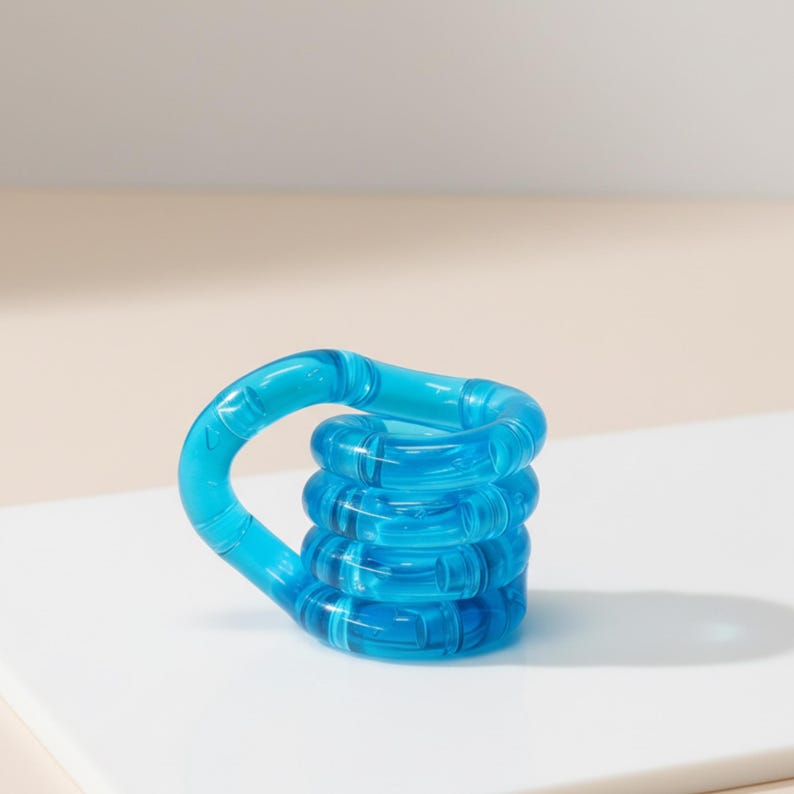 Pode incluir: Um brinquedo fidget azul transparente com uma al&ccedil;a e um corpo empilhado em forma de anel. O brinquedo &eacute; feito de um material liso e brilhante e fica em uma superf&iacute;cie branca. O design &eacute; simples e moderno, com foco na cor e na forma.