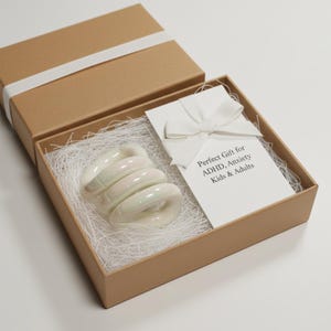 May include: A brown gift box tied with a white ribbon, containing a white, iridescent, spiral-shaped object on white paper. A card with a white bow reads "Perfect Gift for ADHD, Anxiety Kids & Adults."