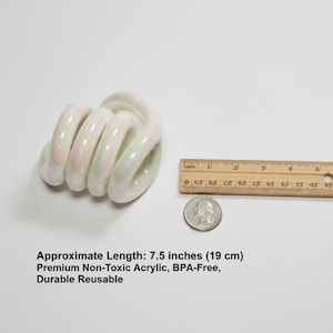 May include: A white and iridescent acrylic toy, approximately 19 cm long, with a spiral design. The toy has subtle pink and green hues. A ruler and a coin are next to the toy for size comparison.