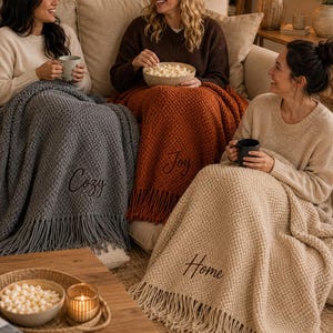 May include: Three textured knit throw blankets with fringe edges, each featuring embroidered text. One grey blanket displays the word Cozy, one rust-coloured blanket features the word Joy, and one cream-coloured blanket shows the word Home.