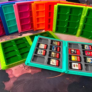 Puede incluir: Una colección de estuches de plástico para guardar cartuchos de juegos en colores vibrantes como morado, azul, rosa, naranja, rojo y verde. Un estuche turquesa abierto en primer plano contiene varios cartuchos de juego rectangulares.
