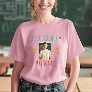 Op de afbeelding: Lichtroze T-shirt met de tekst "BEST TEACHER" in lichtblauw, een hartsymbool en de naam "MRS JONES" in rood. Het T-shirt heeft een foto van een vrouw en is een ronde hals.