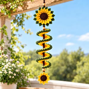 May include: A hanging crochet decoration featuring a large sunflower at the top and a smaller sunflower at the bottom, connected by a central spiral element in yellow and dark green yarn, accented with light wooden beads on a brown cord.