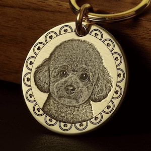 May include: A gold-colored metal keychain with a circular pendant featuring an engraved image of a poodle. The pendant is surrounded by a decorative border with heart-shaped accents. The keychain ring is also gold.