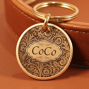 Op de afbeelding: Een ronde, goudkleurige hondenpenning met de naam "CoCo" gegraveerd in een decoratief kader. De penning heeft een sierlijk, wervelend bloemmotief en is bevestigd aan een gouden ring. De achtergrond is een bruin lederen oppervlak.