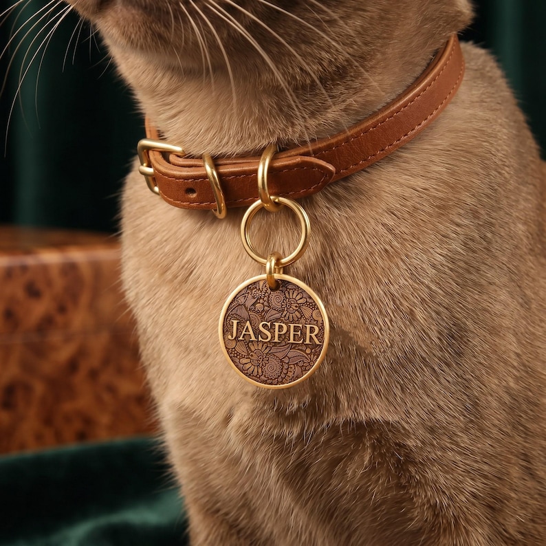 May include: A tan leather pet collar featuring a gold-toned metal buckle and a circular gold-toned pendant. The pendant displays an intricate floral pattern with the name JASPER engraved in the center, attached by a gold-toned metal ring.