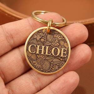 May include: A round gold-toned metal keychain tag featuring an intricate, engraved floral and swirling pattern across the surface. The name CHLOE is embossed in large, capitalized serif letters across the center of the circular pendant.