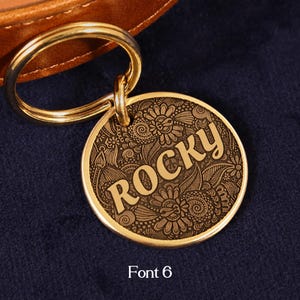 May include: A round gold-toned metal pet ID tag featuring an intricate engraved floral pattern and the name ROCKY in a bold font. The tag is attached to a gold-toned split ring, which is connected to a brown leather collar strap.